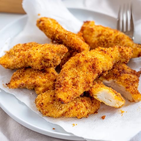 Homemade crispy fried chicken tenders stacked on a wooden board beside a small bowl of ranch dressing.  